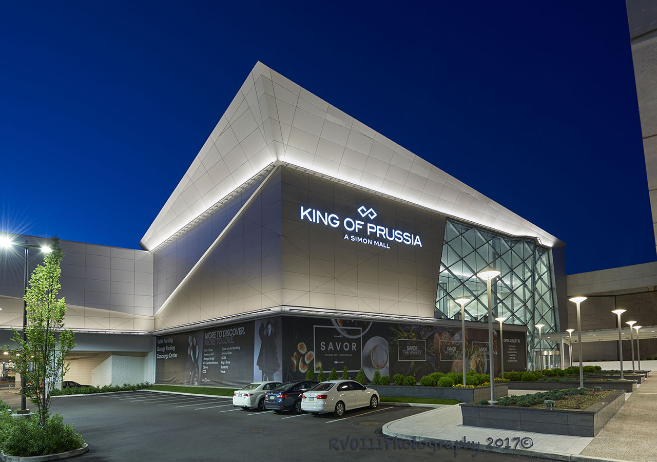 An nighttime exterior of the King Of Prussia Mall with a light up sign that says King of Prussia and a parking lot in the front.