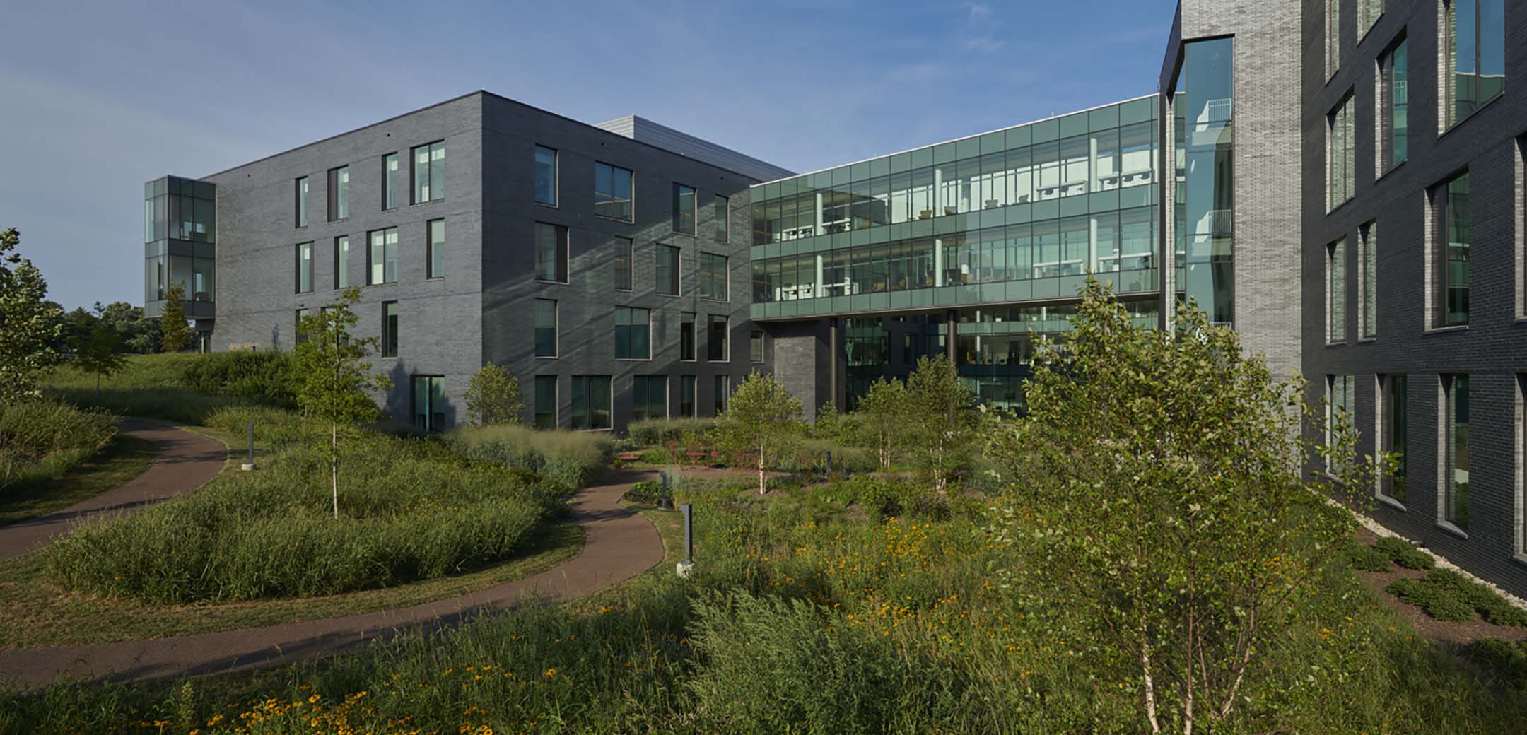 A back view of the Advanced Outpatient Care Center building showcasing the glass connecting bridge and back garden path with bushes and flowers.
