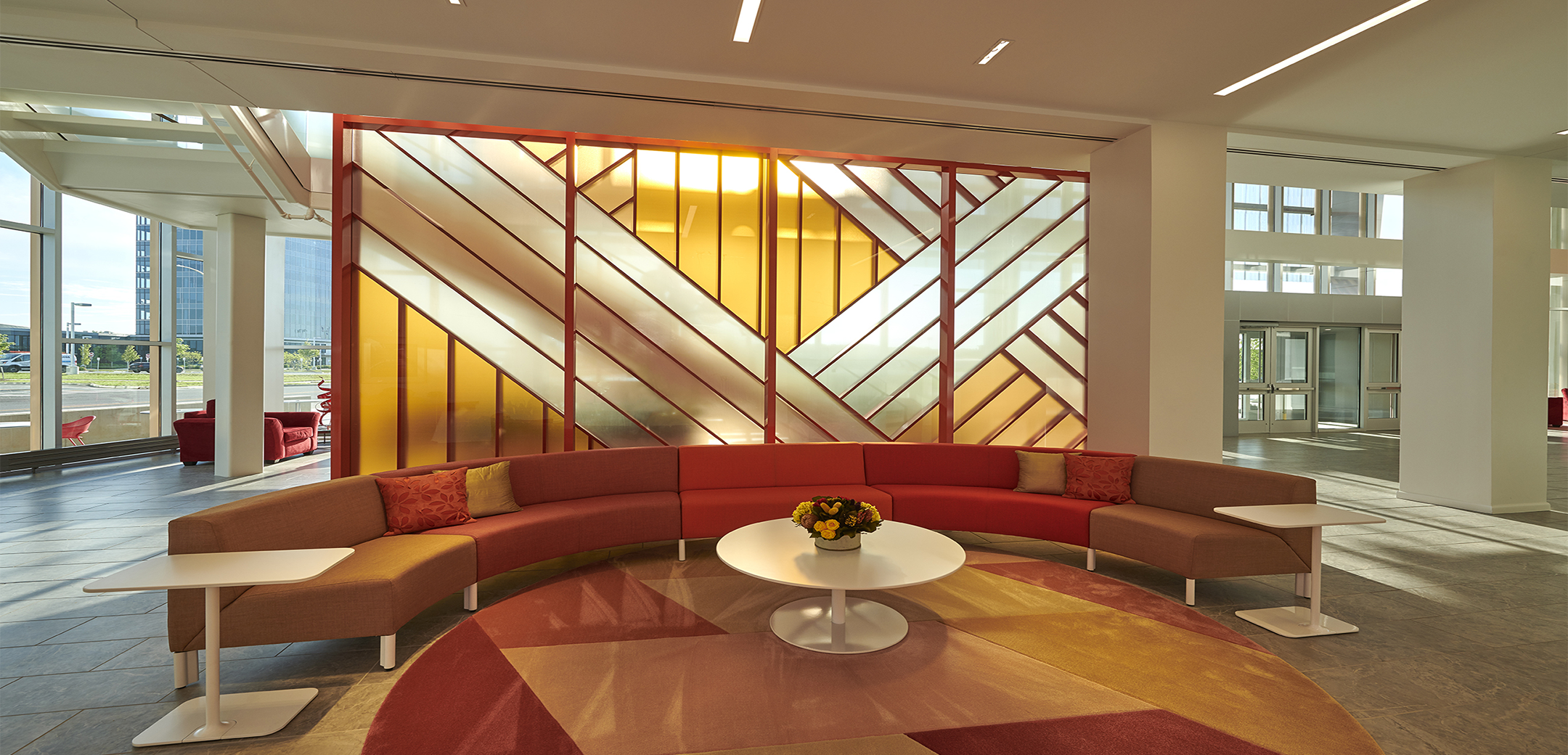 A sitting area in the Chemours Discovery Hub lobby with an orange sofa, circle shaped coffee table, and end tables in front of a yellow glass panel wall.