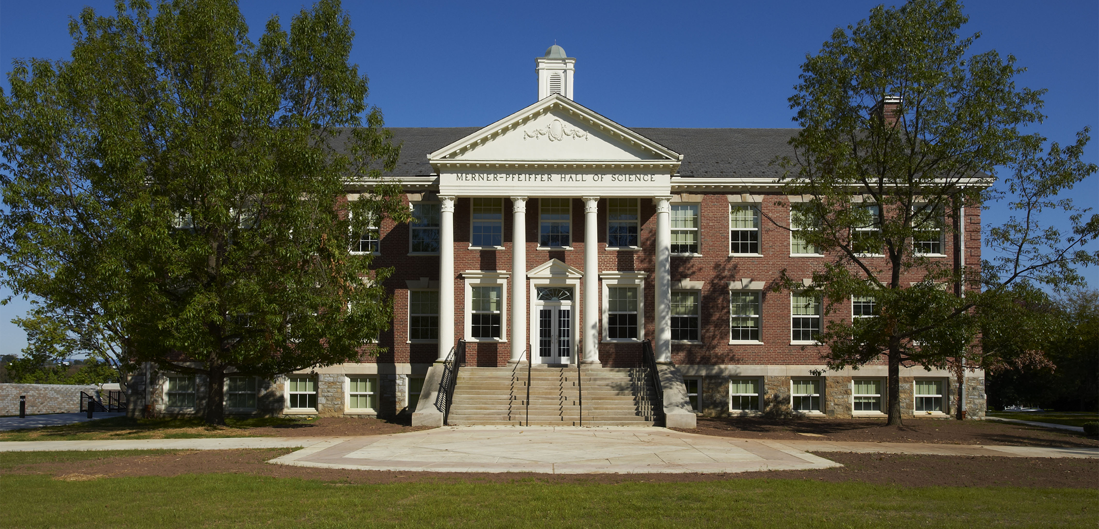 An exterior view of the Merner Pfeiffer Hall of Science building showcasing the front brick design, white columns, staircase leading up to the entrance and a front lawn in the foreground.
