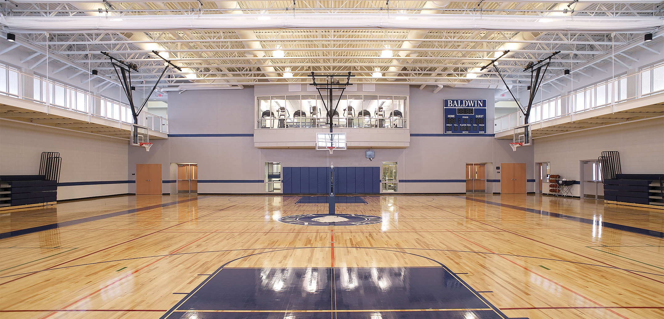 An interior view of the Baldwin School gym, showcasing the lacquered wood flooring, retractable basketball hoops, high ceiling and an elevated gym window in the background.
