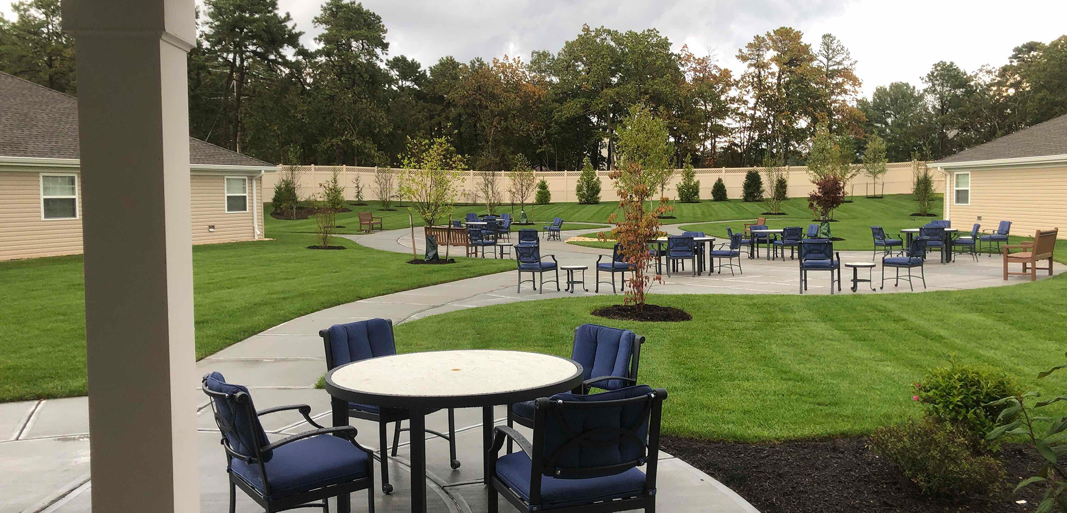 A close up exterior of the backyard of Artis Senior Living Brick with chairs and tables, a paved sidewalk going around the property, and landscaping.
