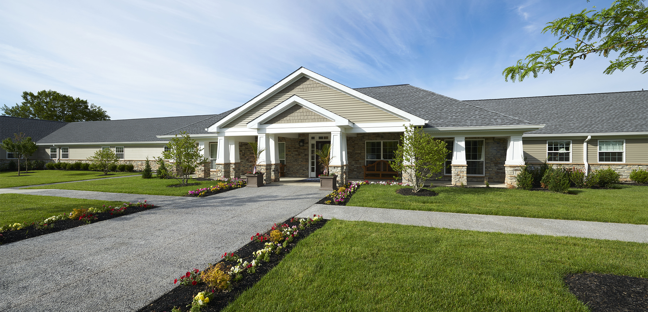 An exterior view of the Artis Senior Living single floor stone brick design building showcasing the overhang triangular design with a grass front lawn and sidewalk going across it.