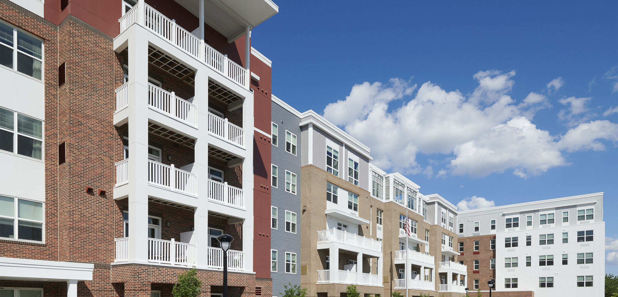 An angled view of the Brightview Devon mixed red brick and panel design building showcasing the white balconies, front entrances and two sided parking lot.