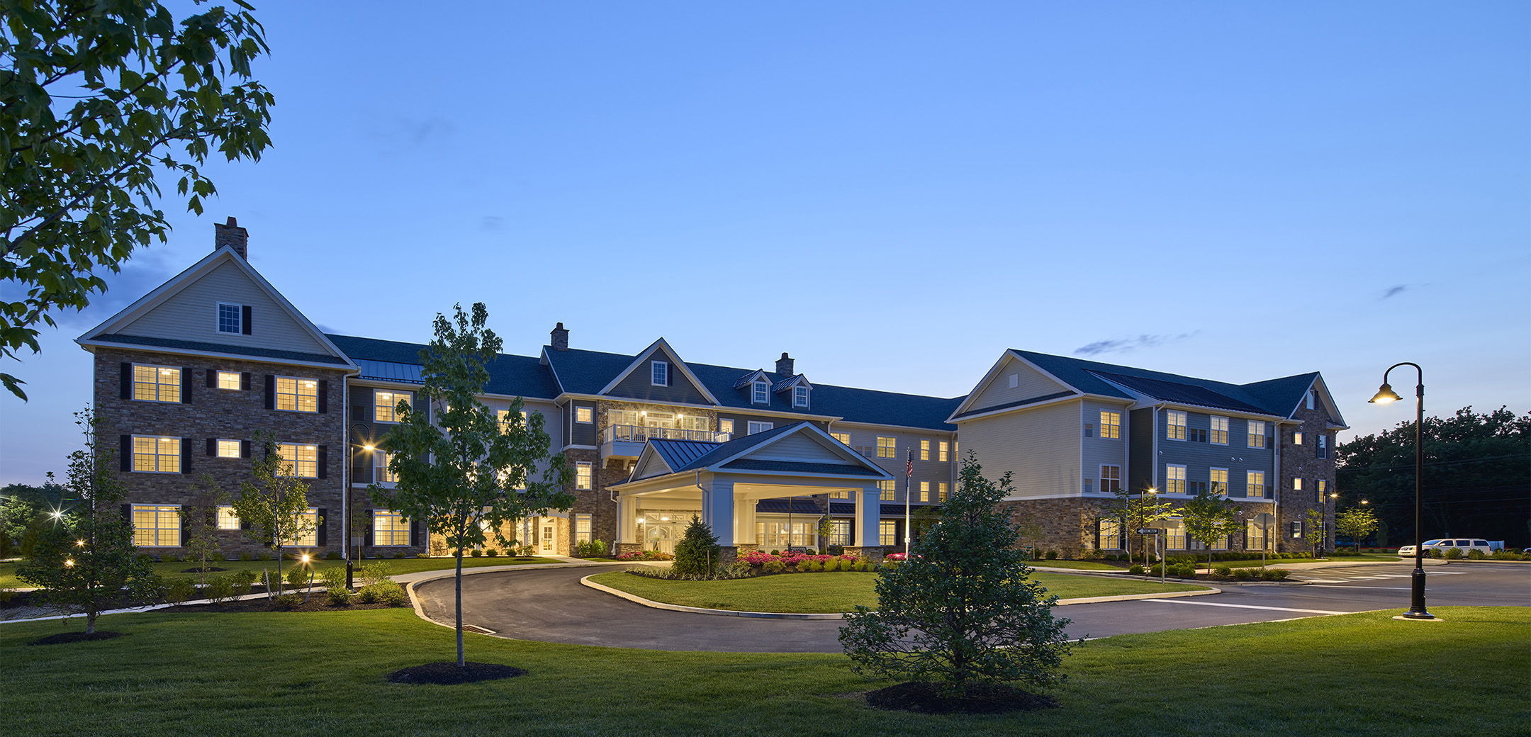 An exterior nighttime image of the Arbor Terrace Exton stone brick, white wood accents and grey shingle roofing with a driveway, side parking lot and grass lawn with trees in the foreground.