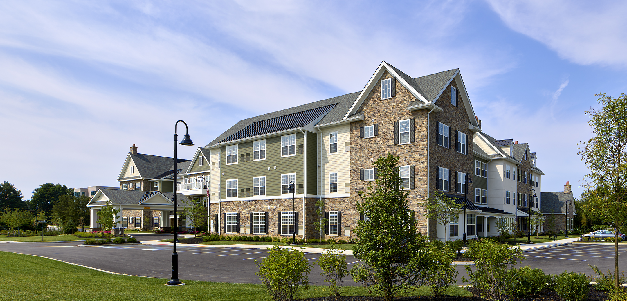 An angled exterior view of the Arbor Terrace Exton showcasing the side parking lot, front overhang entrance with a driveway and green grass in the foreground.