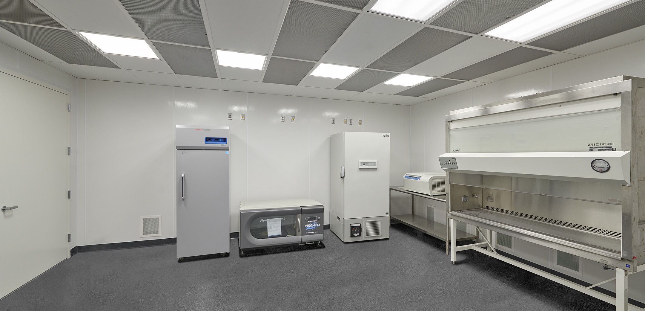 A view of the Charles River Clean Room with panel lighting, gray flooring and white walls, showcasing the test sample fridges and storage units as well as a fume chamber along the wall.