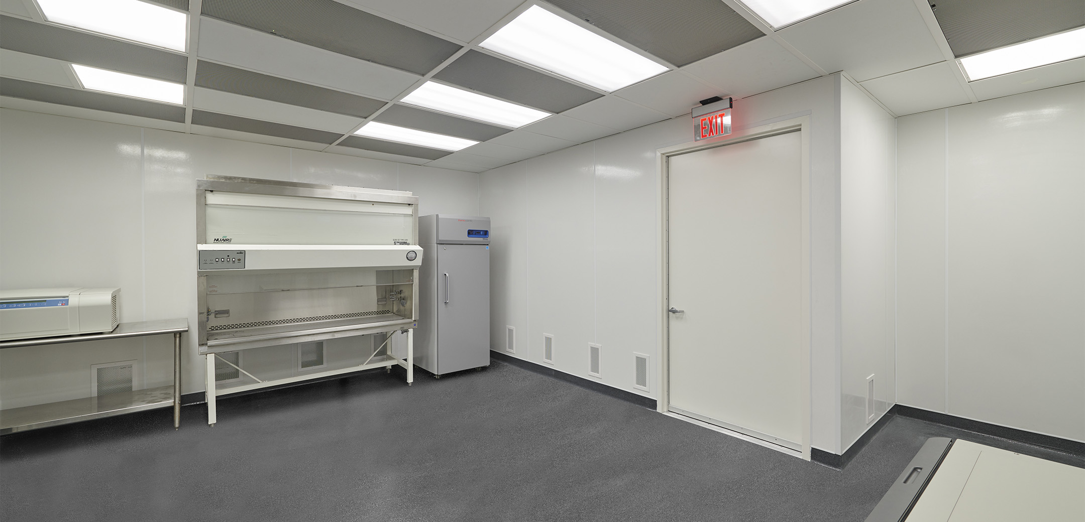 A view of the Charles River Clean Room with panel lighting, gray flooring and white walls, showcasing the large exit door and sanitization equipment along the wall.