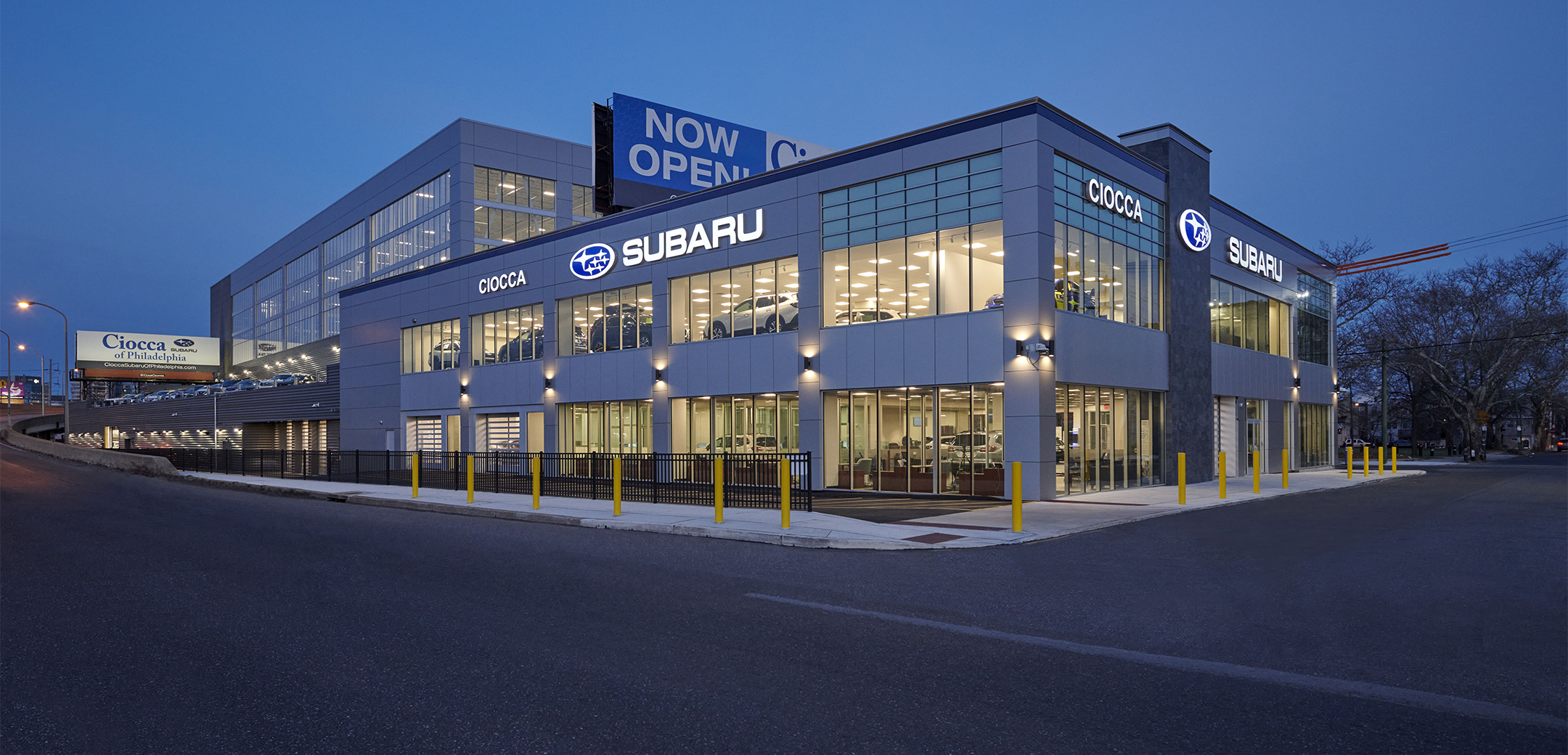 An exterior night view of the Ciocca Subaru two story, large glass window design building showcasing the cars, ``Subaru`` logo, driveway and highway entrance road on the side.