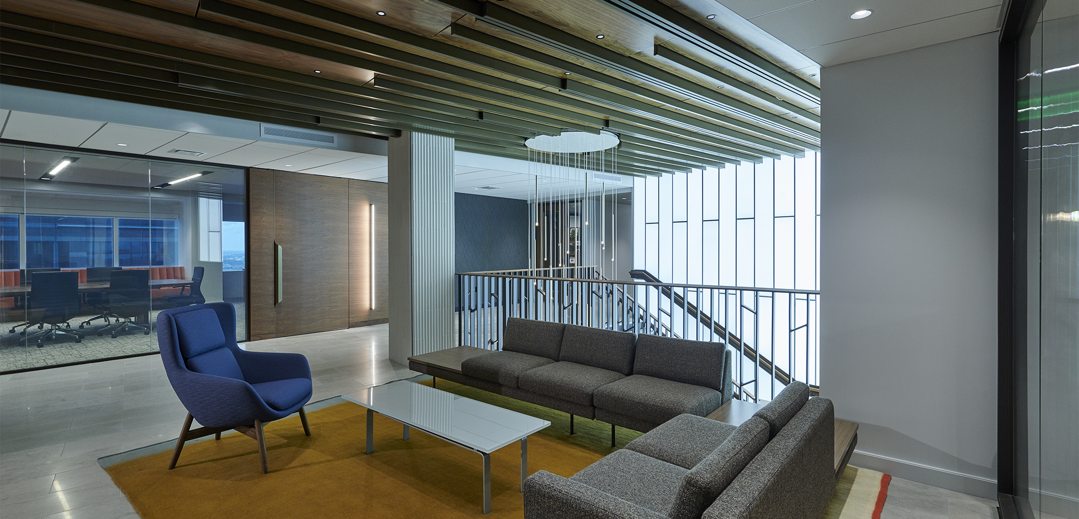 An interior view of the Cohen Seglias showcasing the glass coffee table, two gray couches and a blue chair in the foreground, the staircase and meeting room in the background