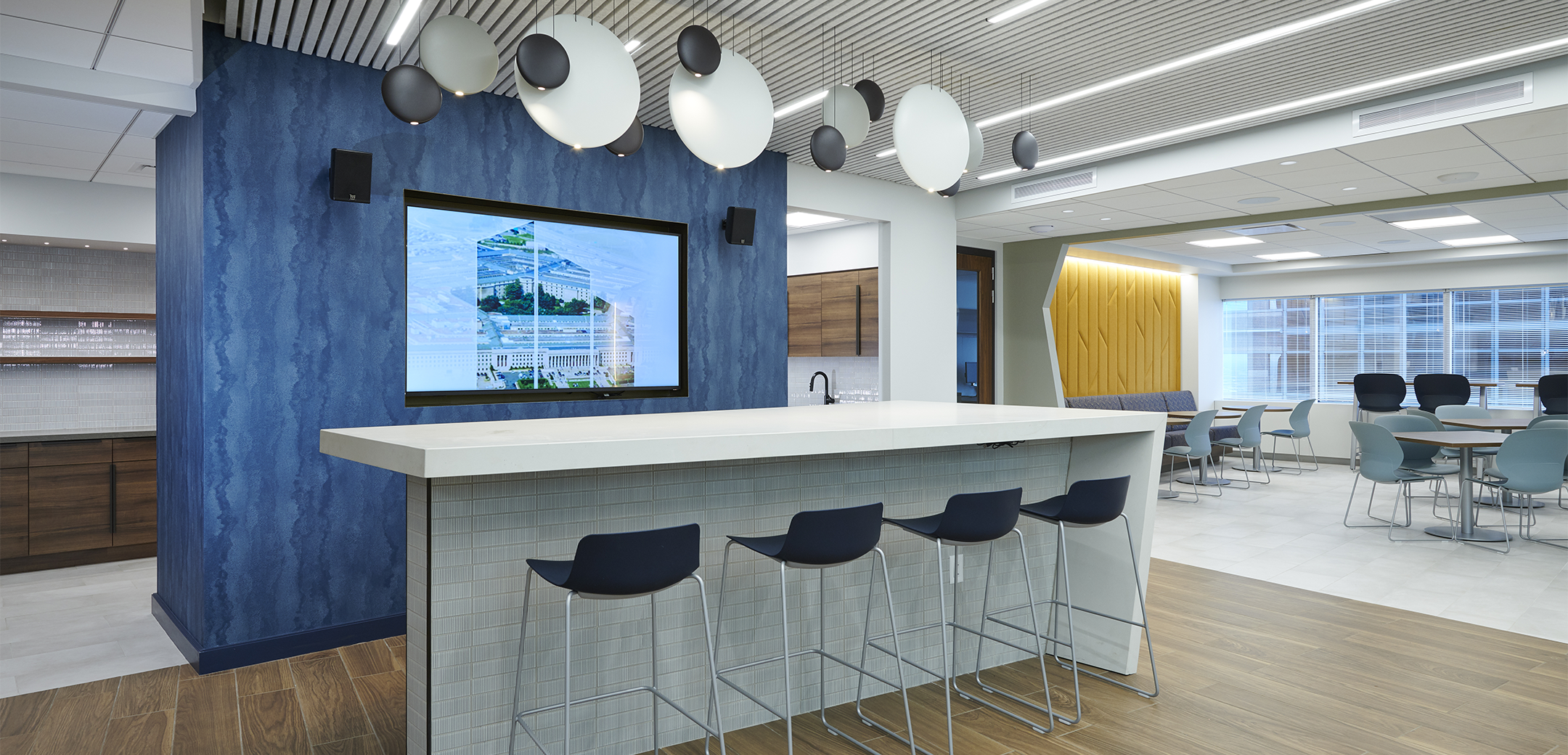 An angled interior view of the Cohen Seglias rest space, showcasing the wooden flooring, blue accent pillar, yellow accent a wall and a centerpiece bar.