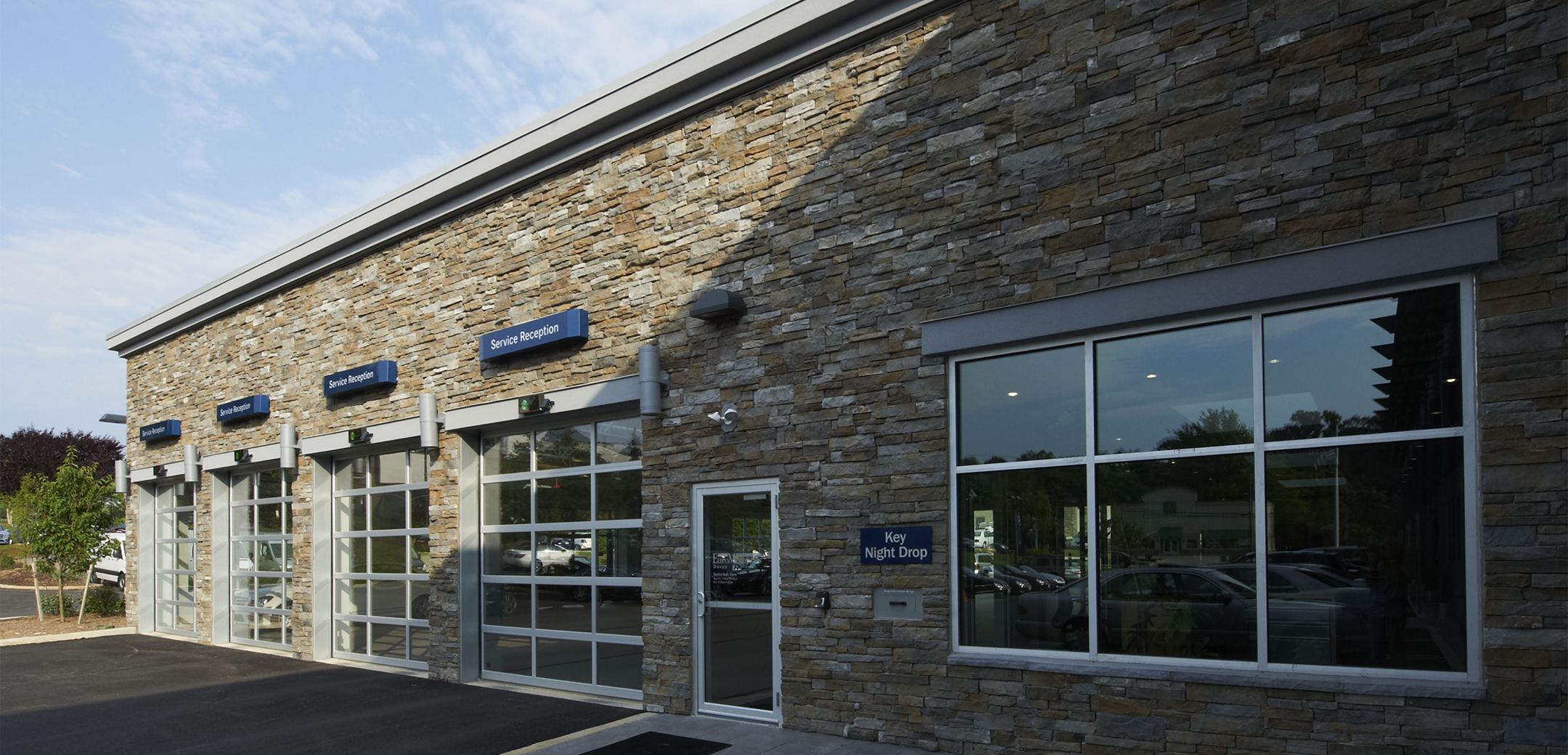 The back side exterior of the Euro Motorcars showcasing the stone brick design with glass shutter garage doors, back exit and a driveway.