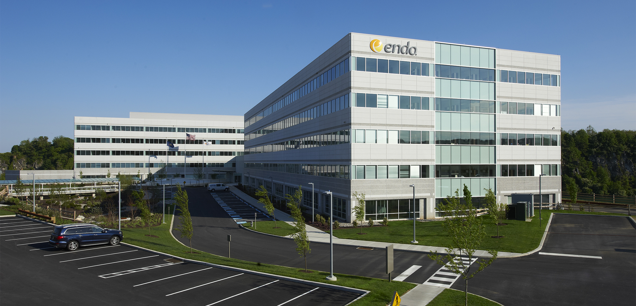 The exterior of a 5-story Endo Pharmaceutical building featuring precast panels and continuous glass window strips showcasing the main front entrance driveway and parking lot.