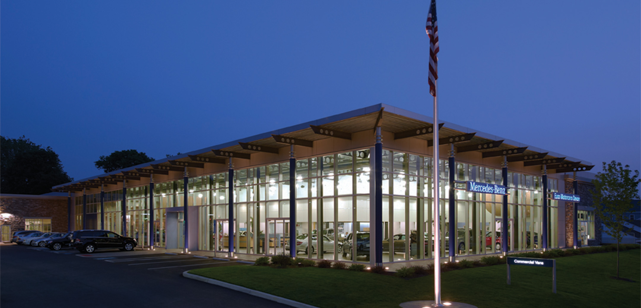 An exterior nighttime view of the Euro Motorcars glass floor to ceiling design building showcasing the cars inside, main parking lot and a flag pole in the foreground with a US flag on it.