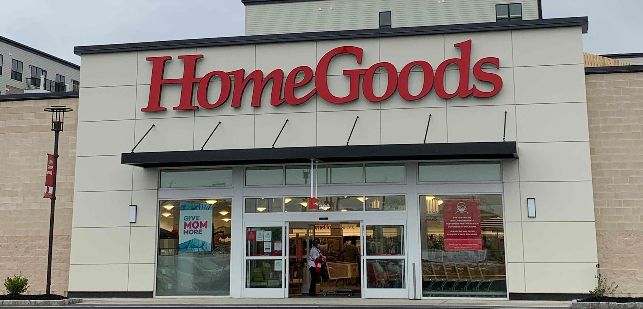 The exterior of the Homegoods building showcasing the brick and concrete slab design with red ``HomeGoods`` signage above the automatic front doors and driveway.