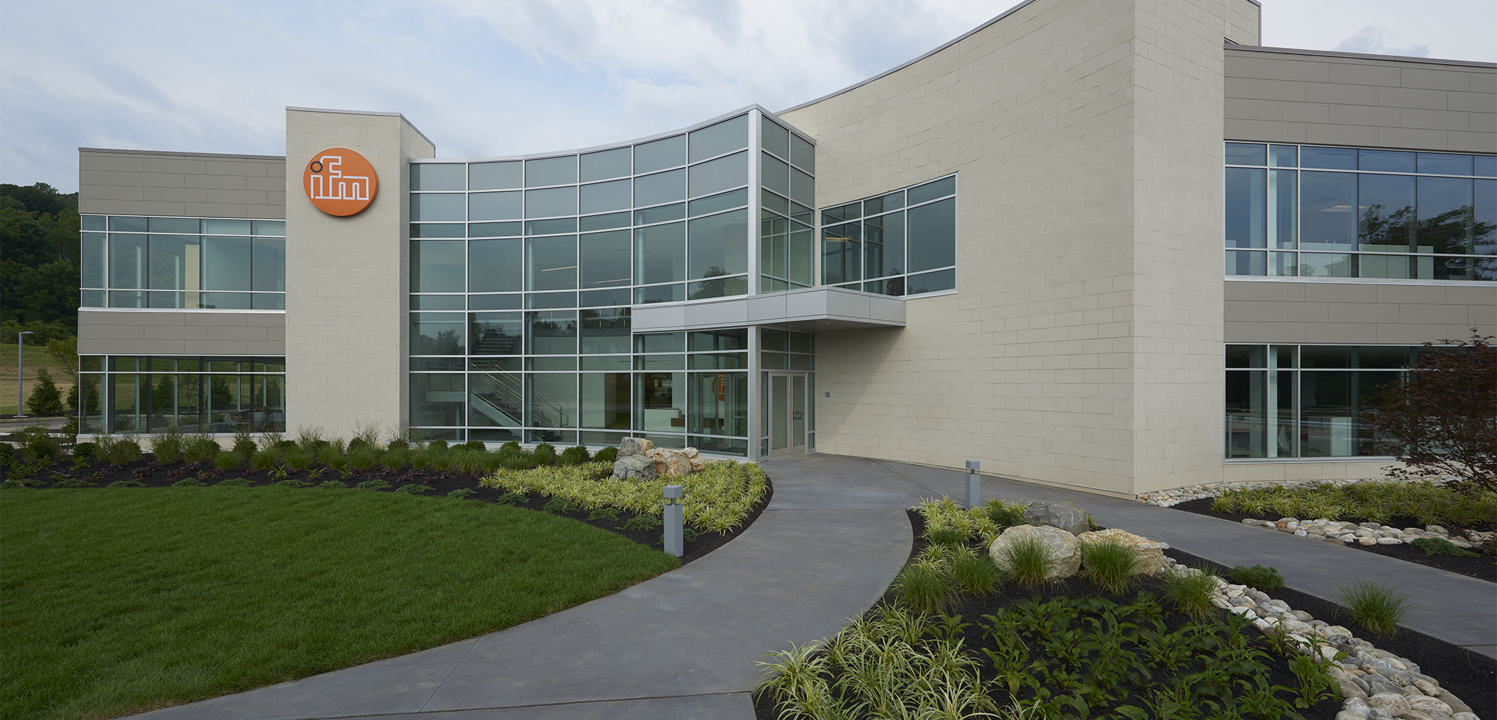 IFM Erector featuring a two story, modern building with a full glass entry way and a concrete panel path cutting through the lawn to it.