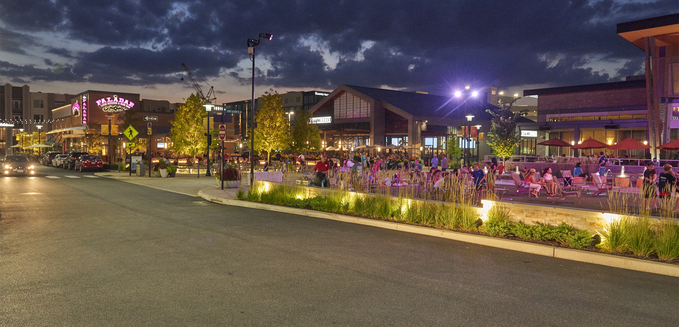 An nighttime exterior view of the KOP Center showcasing the driveway, people sitting on chairs in the center square and grass and tree décor.