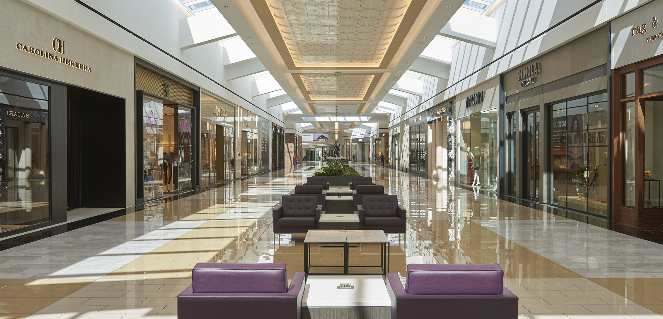 An interior view of the KOP Expansion bridge showcasing the skylights on the ceiling, storefronts on both sides and couch seating areas along the middle of the path going into the background.