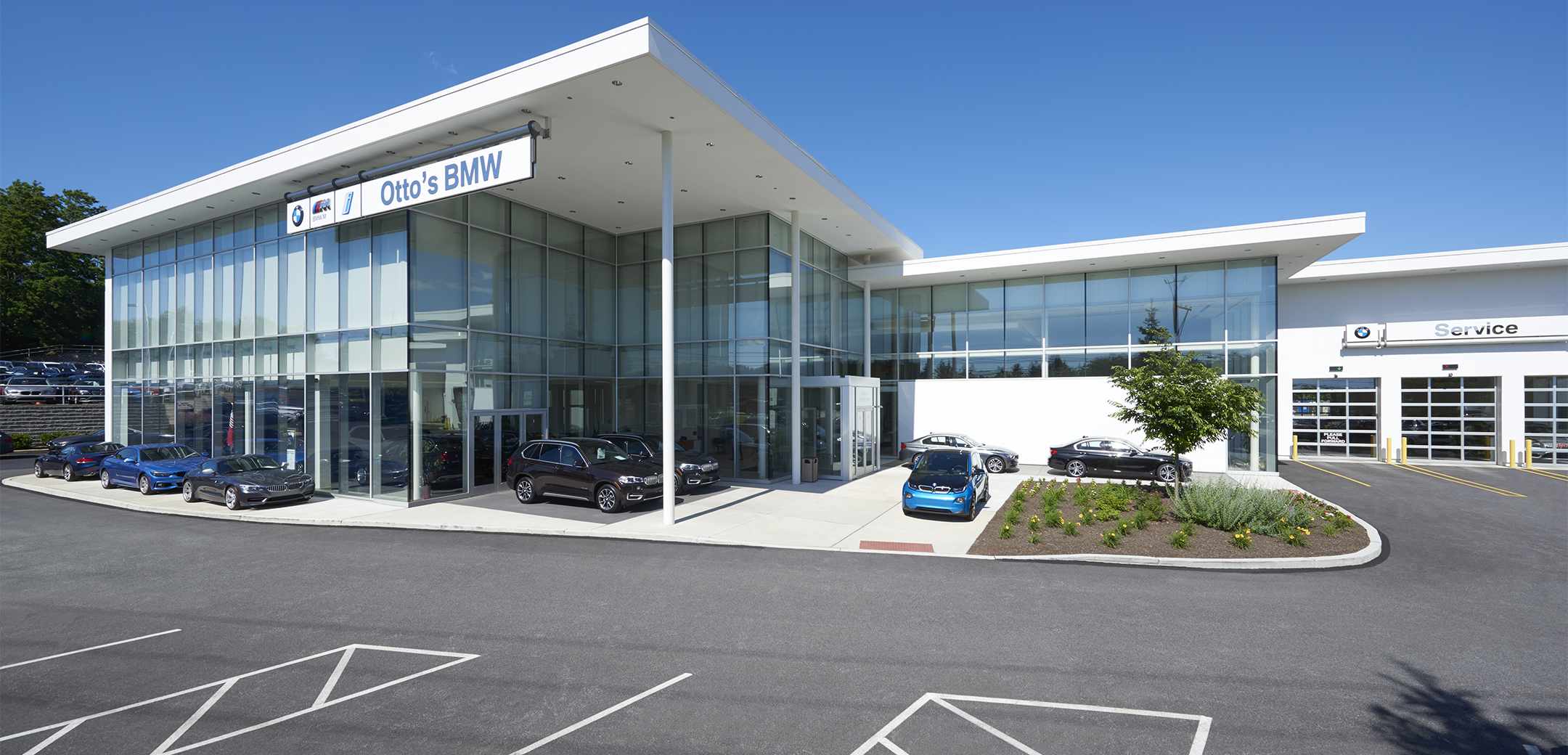 The exterior view of the Ottos BMW building, showcasing the two story, floor to ceiling glass panel design with white support pillars and wall accents with cars placed outside.