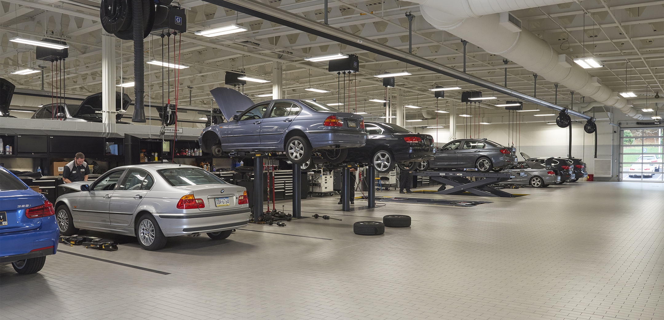 An interior view of the Ottos BMW building with brick tile flooring, tall ceiling, adjustable vacuum on rails, car jacks and a mechanic working on the car.