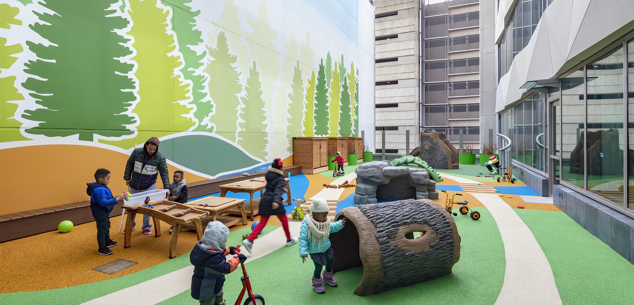 An image of the exterior children's playground with multiple children playing on the playground equipment shaped like trees and rocks.