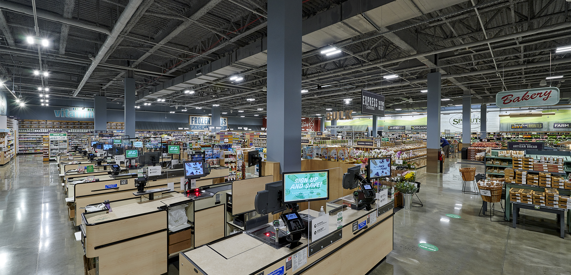 An interior view of the Sprouts Farmers Market showcasing the blue support pillars, tall ceiling, shelves with food and cash registers in the foreground.