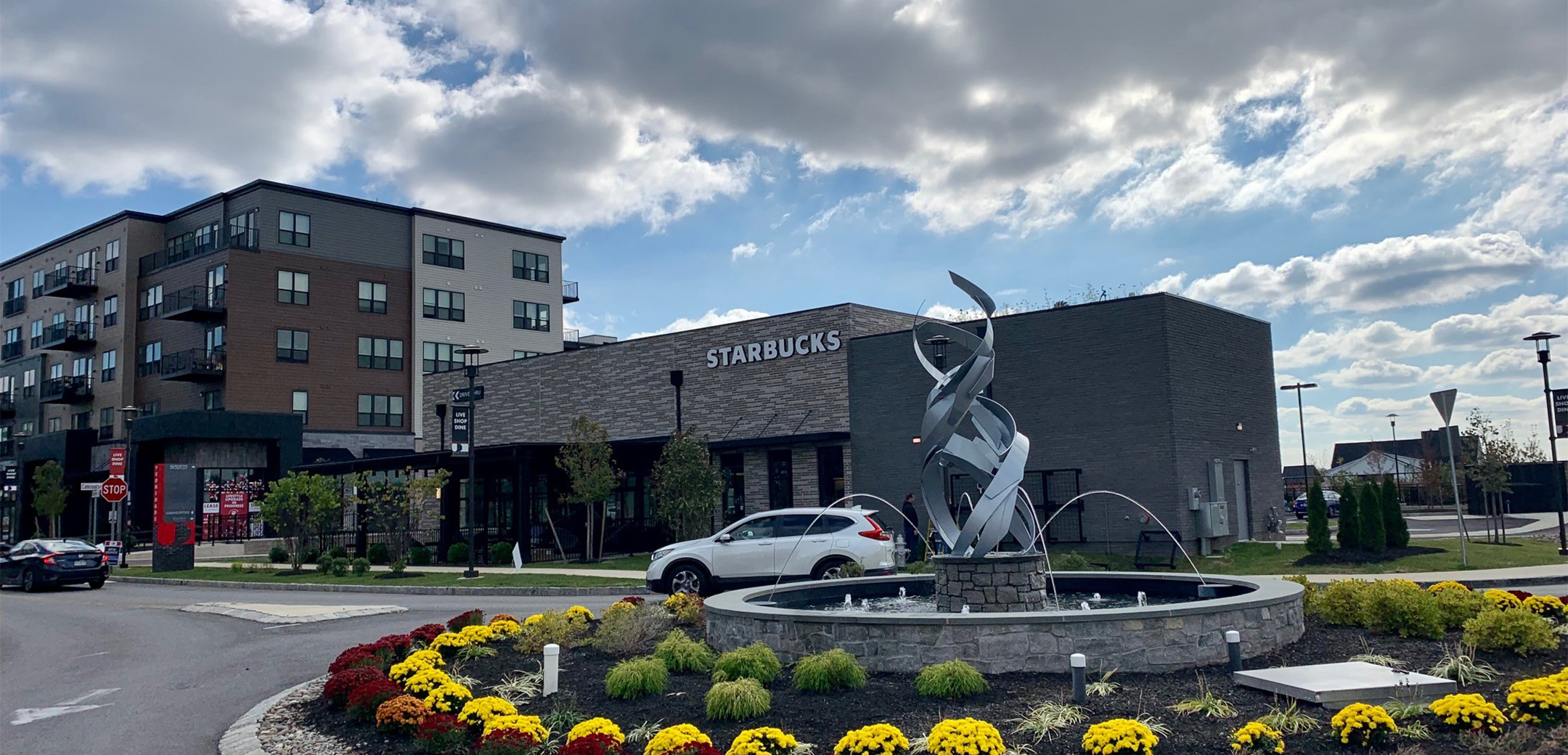 A view of the Starbucks mixed gray brick building showcasing the main street foreground fountain with a metal sculpture in the center and landscaped flowers encircling it.