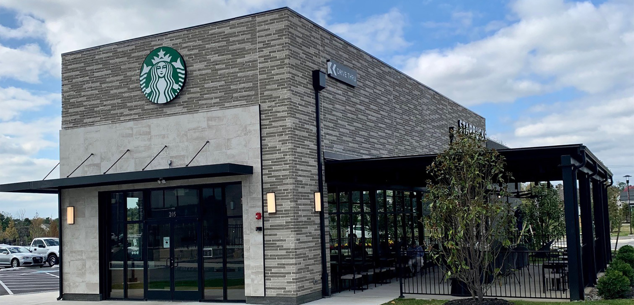 An angled exterior view of the Starbucks front entrance, logo above the overhang glass door and covered outside seating.