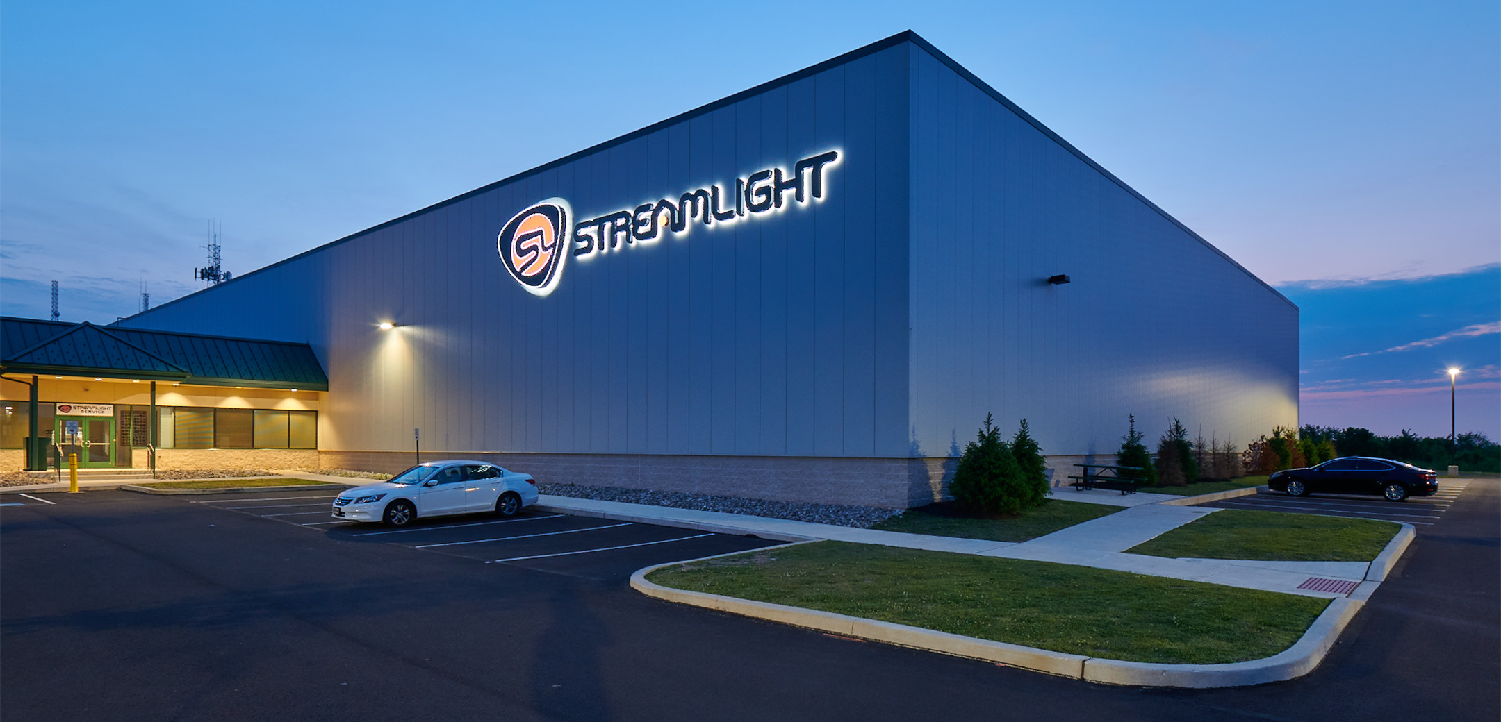 An exterior evening view of the Streamlight warehouse showcasing the lit logo and lettering, front parking and side parking lot.