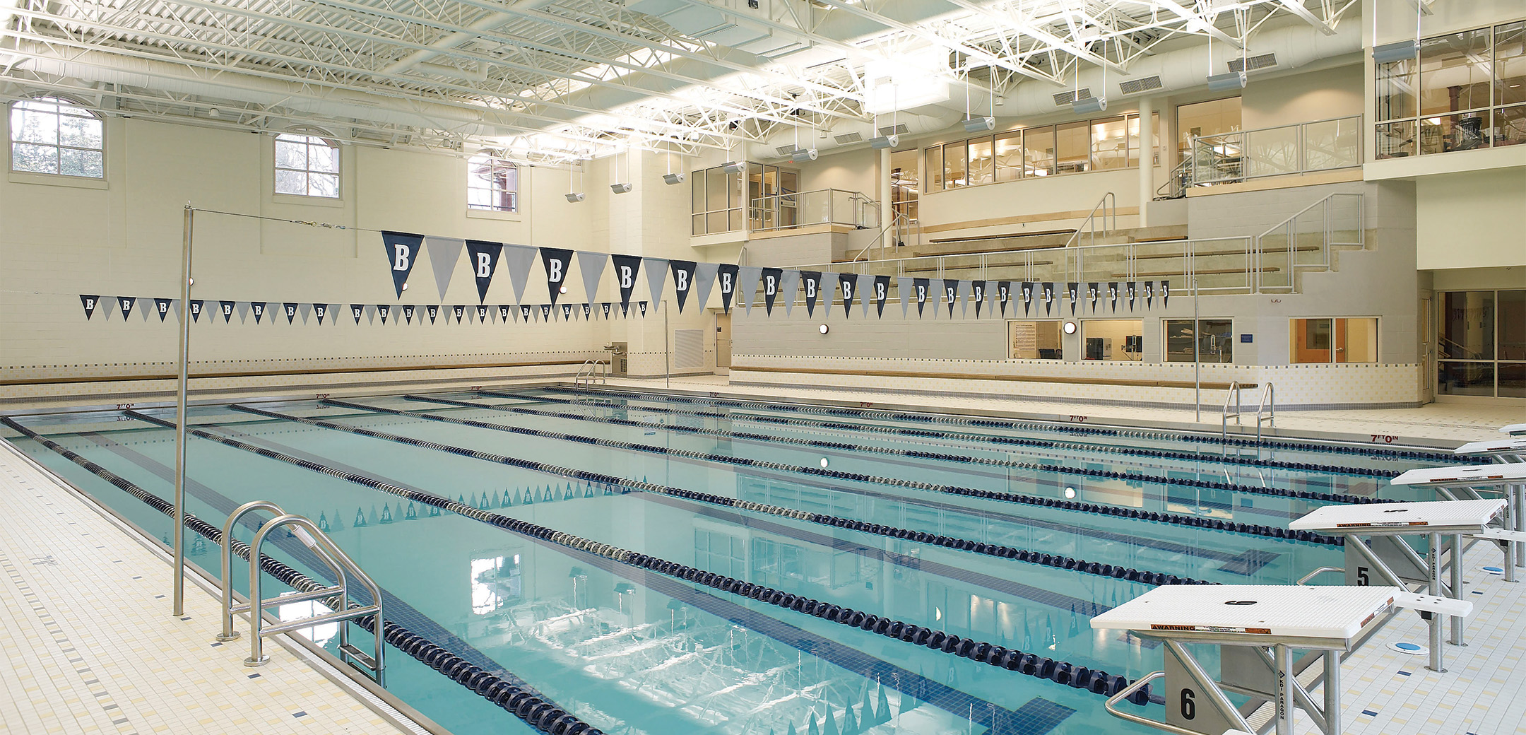 An inner view of the Baldwin School Olympic sized pool with a high well lit ceiling and elevated viewer stands in the background.