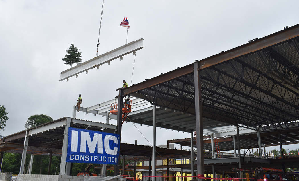 Construction beam with signatures, tree, and American flag being lifted into place in building with large blue IMC Construction banner