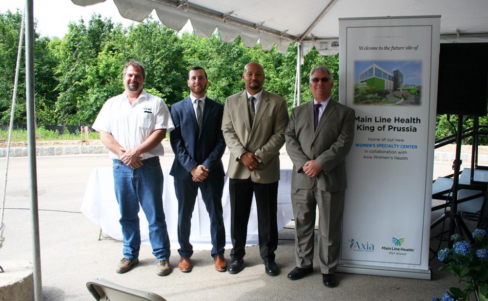 Four IMC Construction members smiling at the camera in front of a banner sign for the new Main Line Health King of Prussia facility.