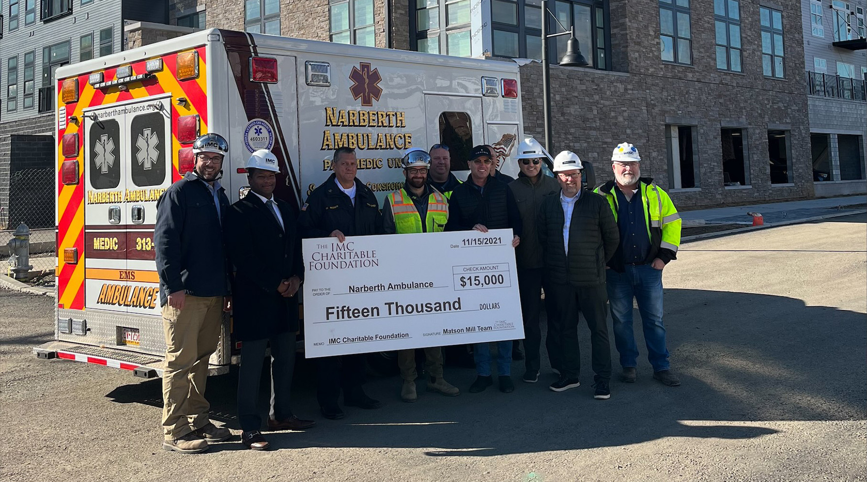 A group of several IMC employees posing with a large donation check in front of an ambulance on a construction site.