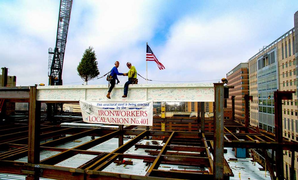 Two workers shaking hands while sitting on a white iron beam and looking at the camera with a sign on the beam that says Ironworkers Local Union