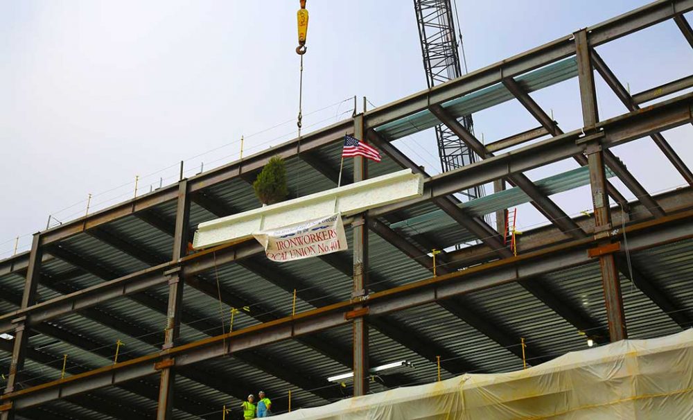 An angled view looking up at a white iron beam with a sign for the Ironworkers Local Union being hoisted by a crane.