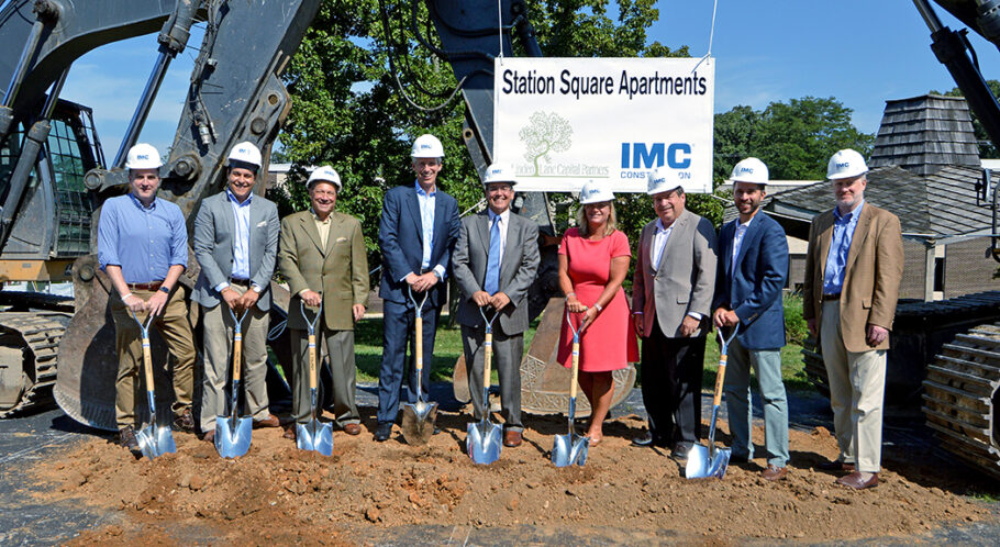 9 members of the IMC Construction team wearing IMC hardhats and holding shovels in front of large machinery with a sign that says "Station Square Apartments" behind them.