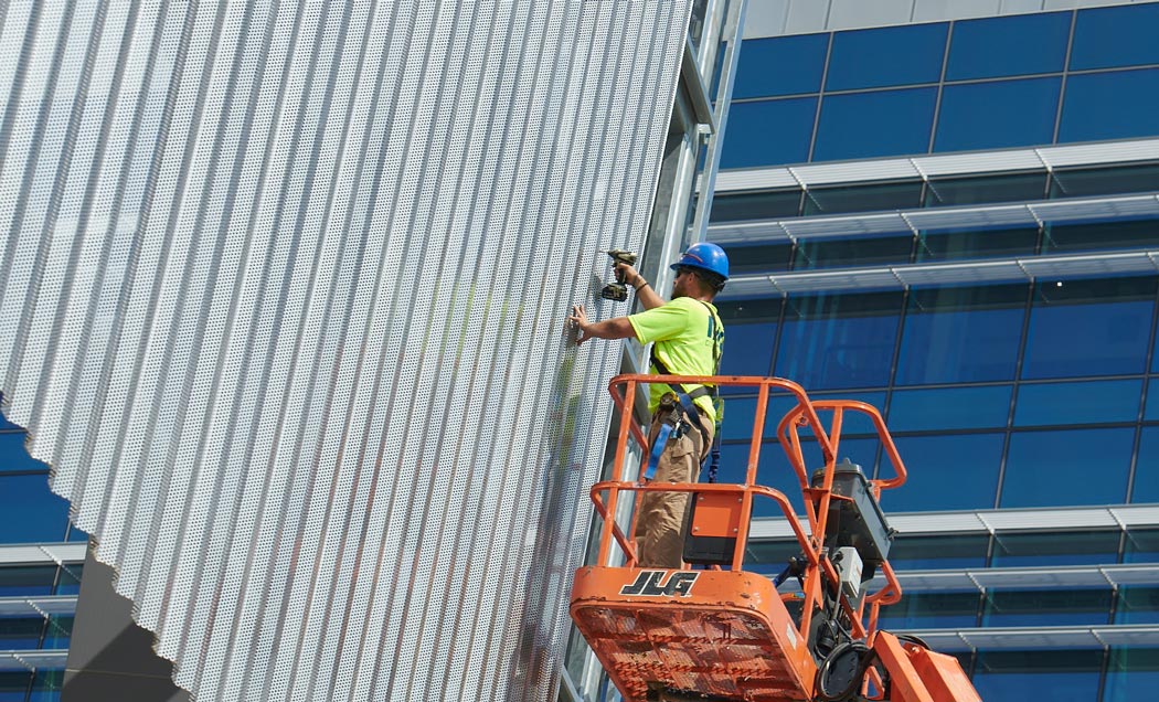 An IMC Construction worker standing in an orange lift operating a power drill on the exterior of a metal building.