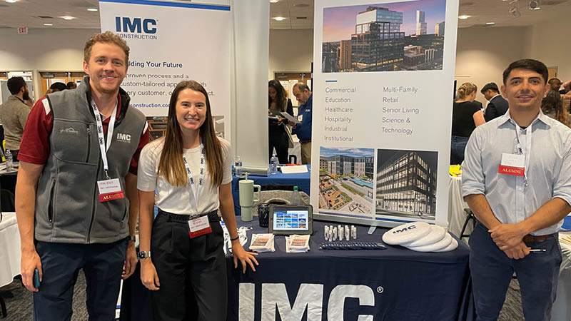 Three IMC employees at Temple University Career Fair 2023 standing in front of table with IMC banners